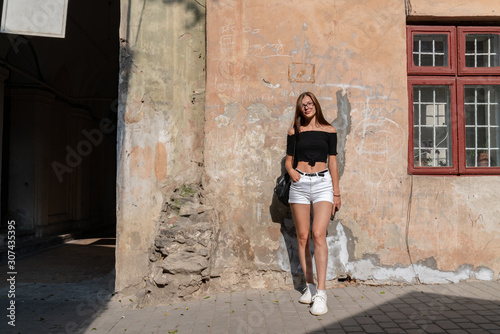 Young attractive brunette girl in glasses, dressed in white shorts and black top, standing near the wall of the old house and looking at the camera