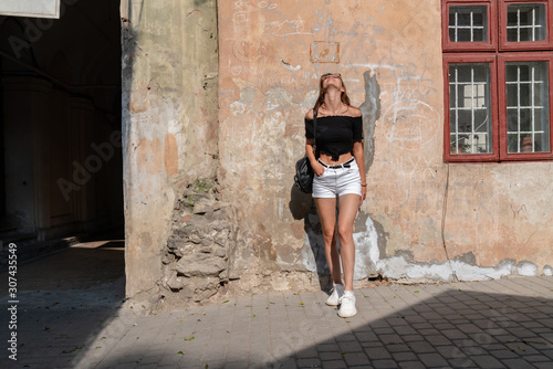 Young attractive brunette girl in glasses, dressed in white shorts and black top, standing near the wall of the old house and looking up