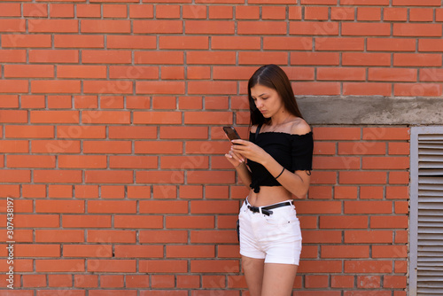 Young attractive brunette girl in glasses, dressed in white shorts and black top, standing near the brick wall, holding phone in her hands