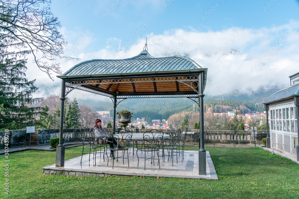 Beautiful wooded place with a pergola and garden furniture, a pretty redhead girl seated in a relaxed attitude. Busteni Romanian village. Carpathian mountains - Transylvanian alps. Beautiful wooded place with a pergola and garden furniture, a pretty redhead girl seated in a relaxed attitude. Busteni Romanian village. Carpathian mountains - Transylvanian alps.