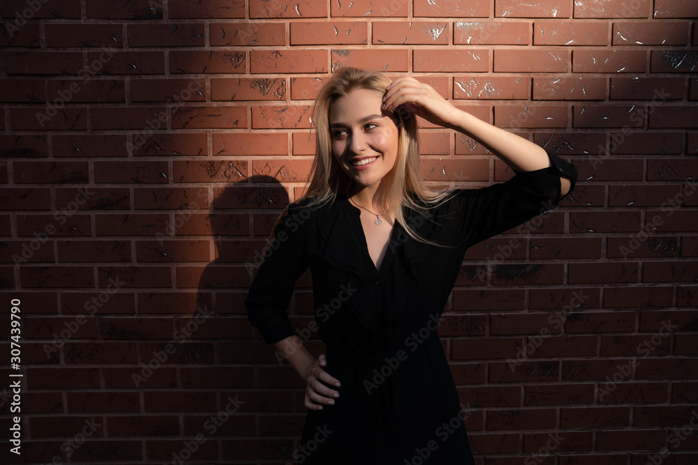 Happy smiling blonde girl holding one hand on her waist, touching her hair with her hand and looking away, red brick wall on the backgroud Happy smiling blonde girl holding one hand on her waist, touching her hair with her hand and looking away, red brick wall on the backgroud
