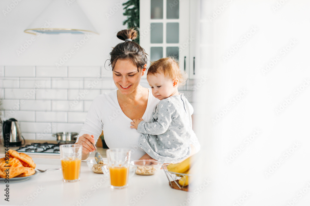 Young woman mom with baby girl on hands cooking breakfast on bright kitchen at home Young woman mom with baby girl on hands cooking breakfast on bright kitchen at home