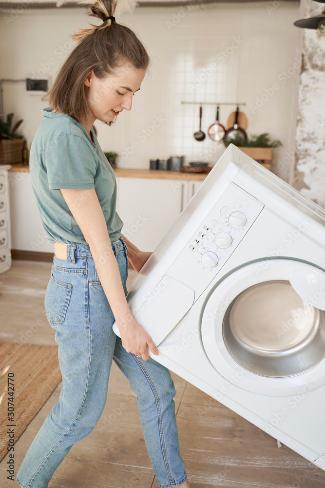 Powerful cheerful young woman trying to carry white washing machine alone in light kitchen Powerful cheerful young woman trying to carry white washing machine alone in light kitchen