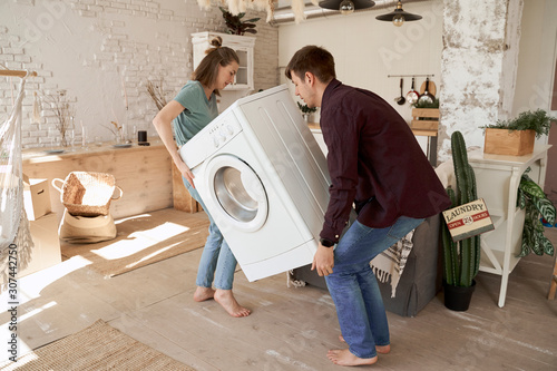 Side view of barefoot content man and woman carrying white washing machine while moving to new house photo