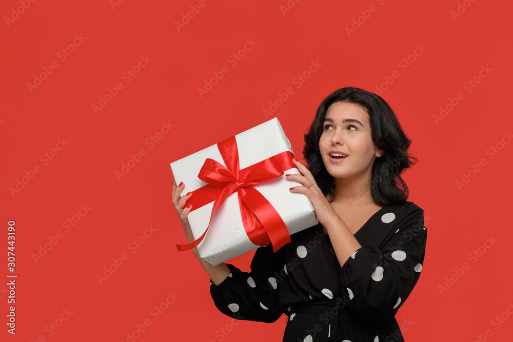 Brunette woman in a black dress smiling holding Christmas giftbox of white color with red ribbon. Brunette woman in a black dress smiling holding Christmas giftbox of white color with red ribbon.