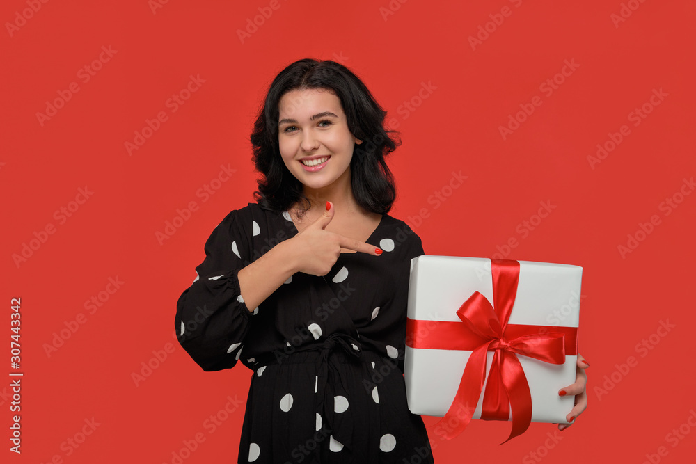 Brunette woman in a black dress smiling holding Christmas giftbox of white color with red ribbon.
