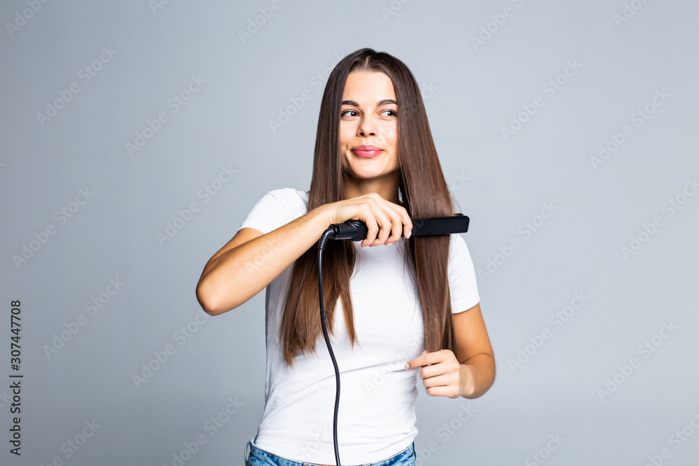 Fototapeta premium Young woman doing hairstyle with hair straightener isolated on gray background