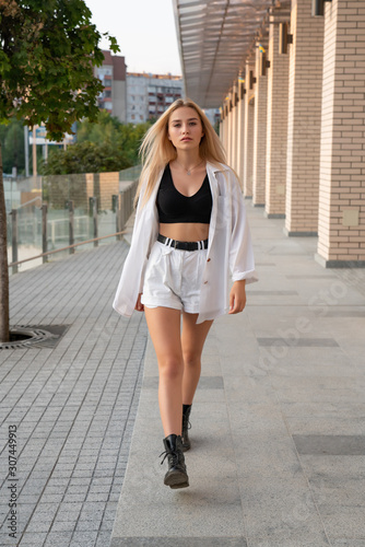 Outdoor street style photo, beautiful blonde girl in white shorts and black boots walks along modern building © Vasya