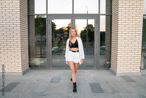 Fashionable stylish blonde girl in white shirt and black boots walks along modern building