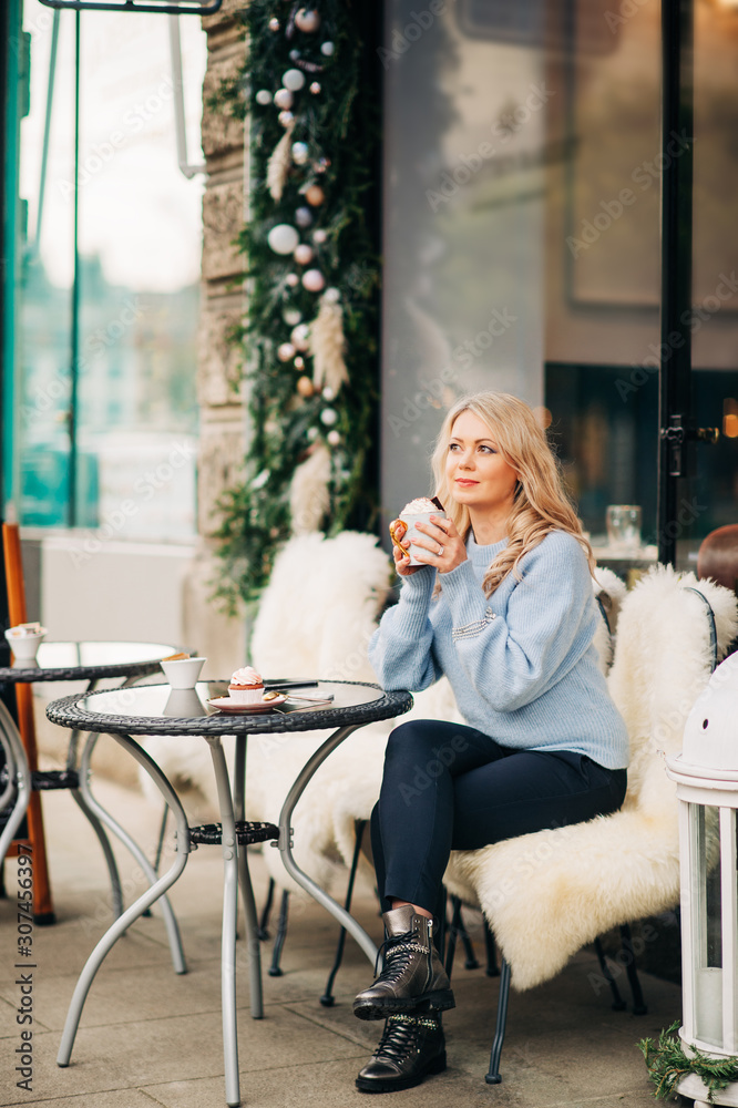 Outdoor portrait of happy beautiful woman relaxing on a terrace of cozy Cafe or Tea Room, holding cup of hot chocolate, wearing blue knitted pullover Outdoor portrait of happy beautiful woman relaxing on a terrace of cozy Cafe or Tea Room, holding cup of hot chocolate, wearing blue knitted pullover