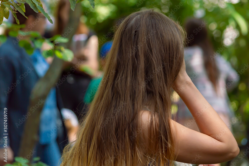 Girl with straight long brown hair straightens her hair. City park, trees, people. Back view. Girl with straight long brown hair straightens her hair. City park, trees, people. Back view.