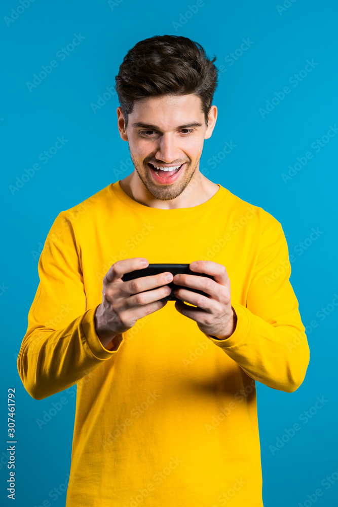Handsome man playing game on smartphone on blue studio wall. Using modern technology - apps, social networks. Handsome man playing game on smartphone on blue studio wall. Using modern technology - apps, social networks.