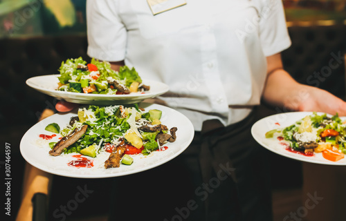 Waitress is holding fresh salad plates in her hand. Woman sets the table at the restaurant. Cafe service for birthday or wedding celebration. Different dishes on the served table.