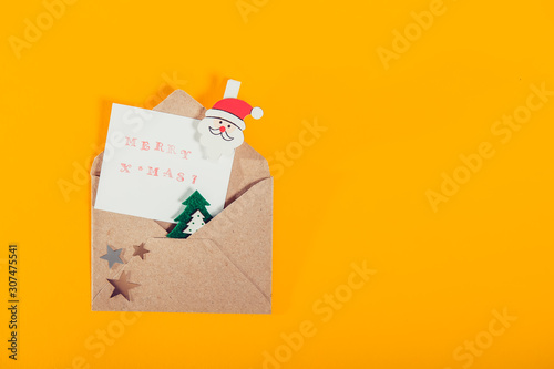 Top view festive composition with handmade stamp greeting card, santa clause pin in craft envelope with felt christmas tree and stars on bright yellow background. Flat lay, top view. Copy space.