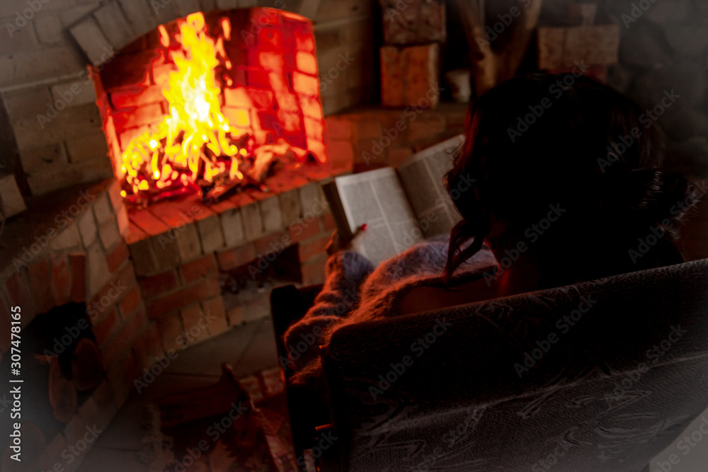 Back view of pregnant woman wearing knitted warm sweater sitting in rocking chair. Female reading the book against the burn fire in brick fireplace at home