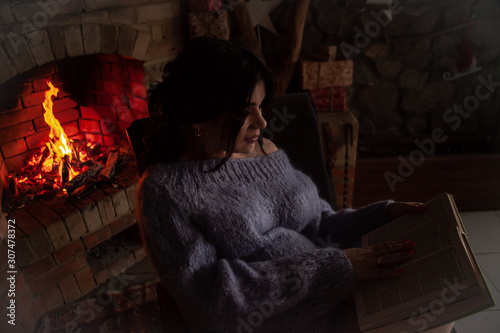 Back view of pregnant woman wearing knitted warm sweater sitting in rocking chair. Female reading the book against the burn fire in brick fireplace at home and holding a cup of tea