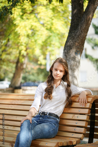  a girl in a white blouse and blue jeans sits on a wooden bench in autumn Park square, miss autumn