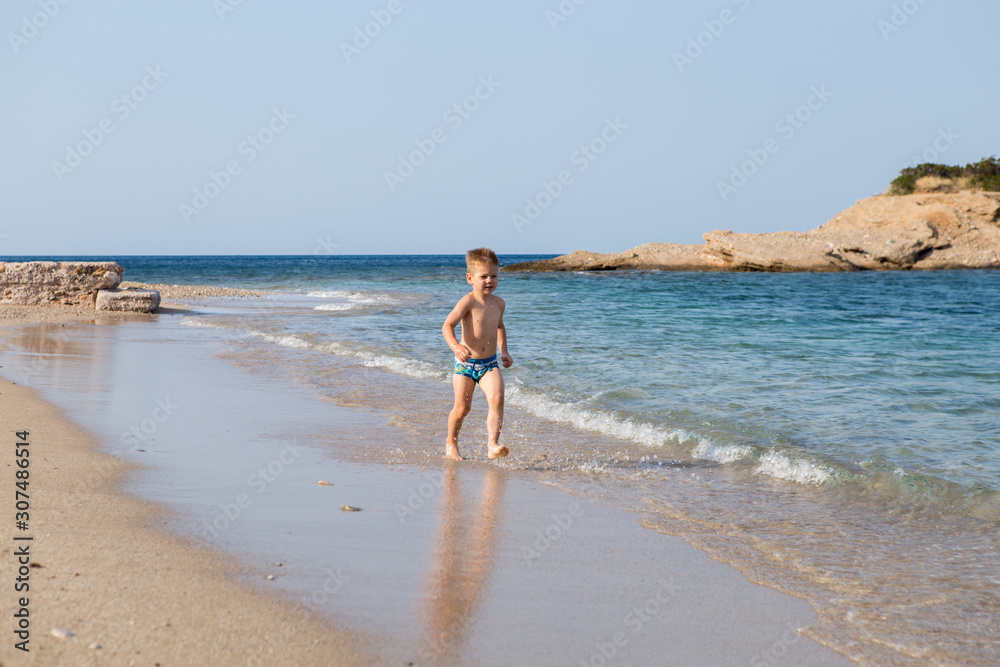 A little cute boy runs along the beach and plays. Happy kid at sea. Vacation at sea with children. Greece A little cute boy runs along the beach and plays. Happy kid at sea. Vacation at sea with children. Greece