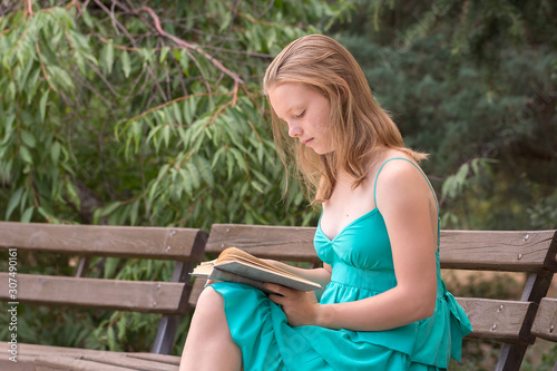 Tableau sur toile A schoolgirl in a turquoise dress is reading a fascinating book on a bench in a summer park