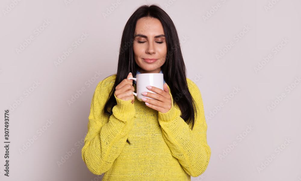 Cozy day. Close-up photo of attractive young woman in a yellow sweater, who is enjoying her hot tea, smiling with her eyes closed.