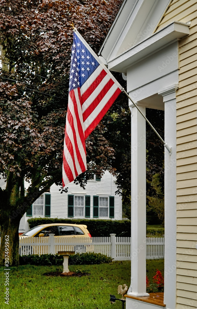 The Stars & Stripes hang ouside a classic Cape Cod house overlooking a lawn with white picket fence and a yellow car driving by.