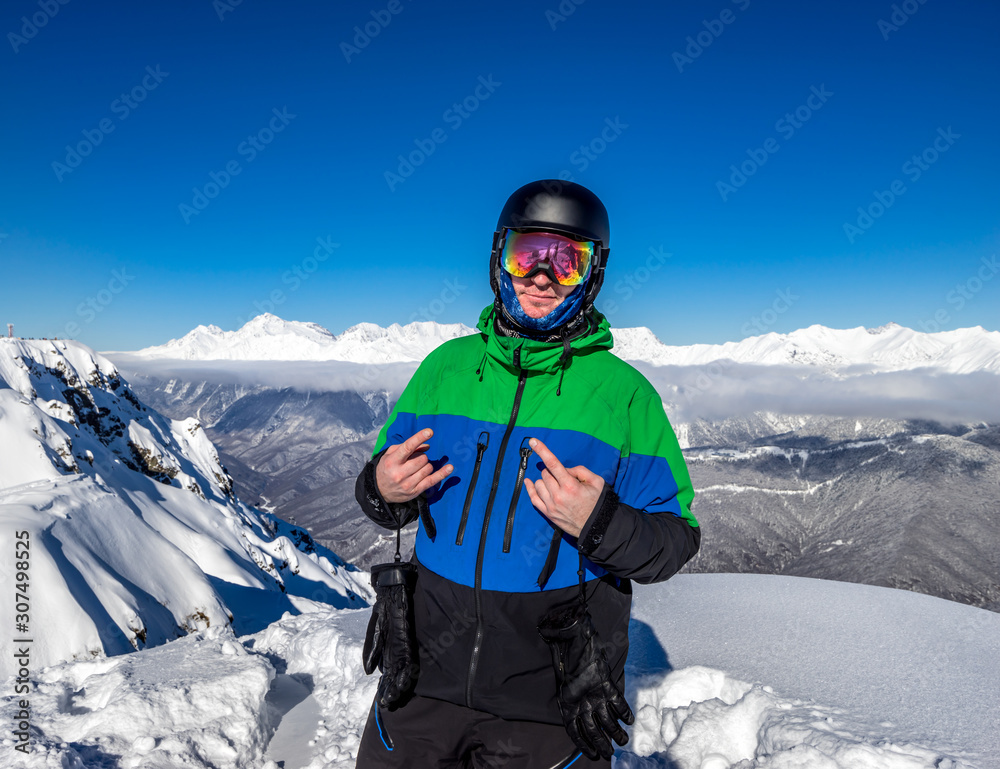 Freerider snowboarder standing on steep slope of mountain peak, showing a sign everything is cool, on background of snowy mountains in ski resort