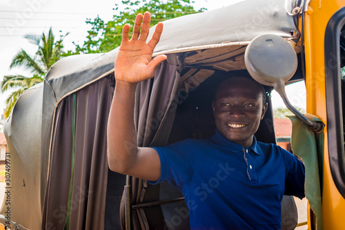 young african man driving a tuk tuk taxi smiling on a very sunny day waving to a passenger photo