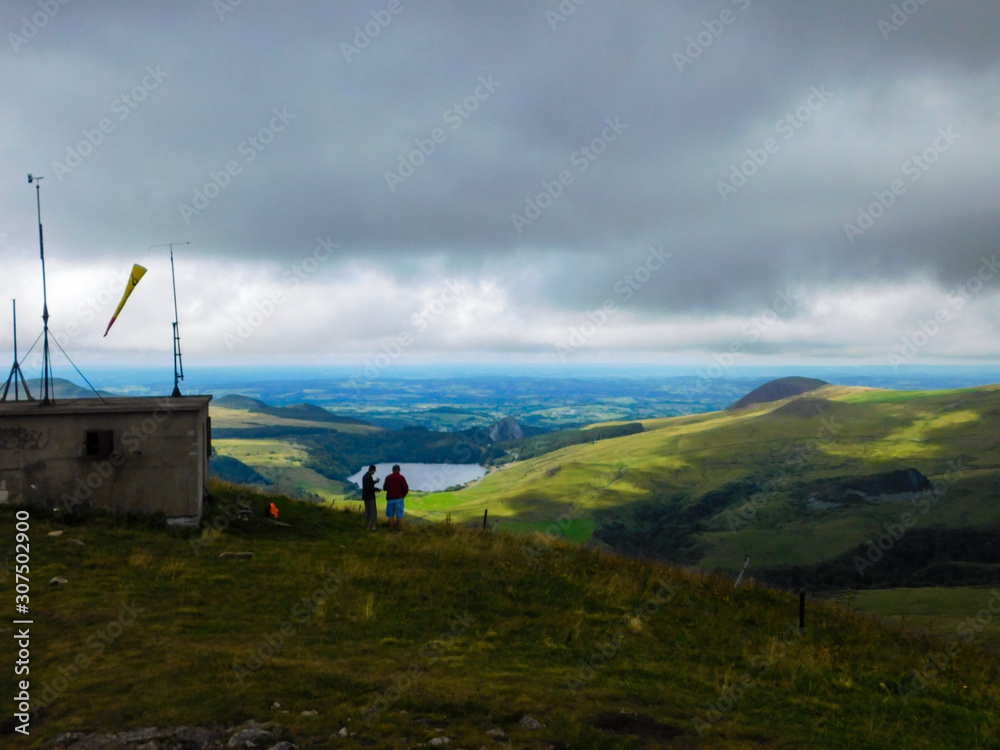 Obraz premium Deux randonneurs au sommet de la montagne dans les volcans d'Auvergne avec vue panoramique sur lac et horizon lointain, belle couche nuageuse grise