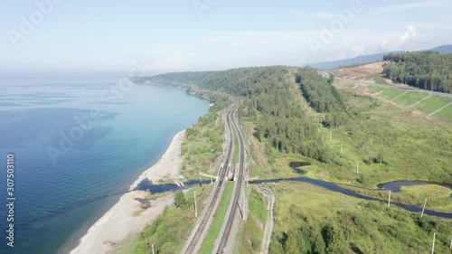 Aerial view of railroad of Trans-Siberian Railway on shore of Lake Baikal with green trees in a sunny summer day. East Siberian Railway in Buryatia, Siberia, Russia