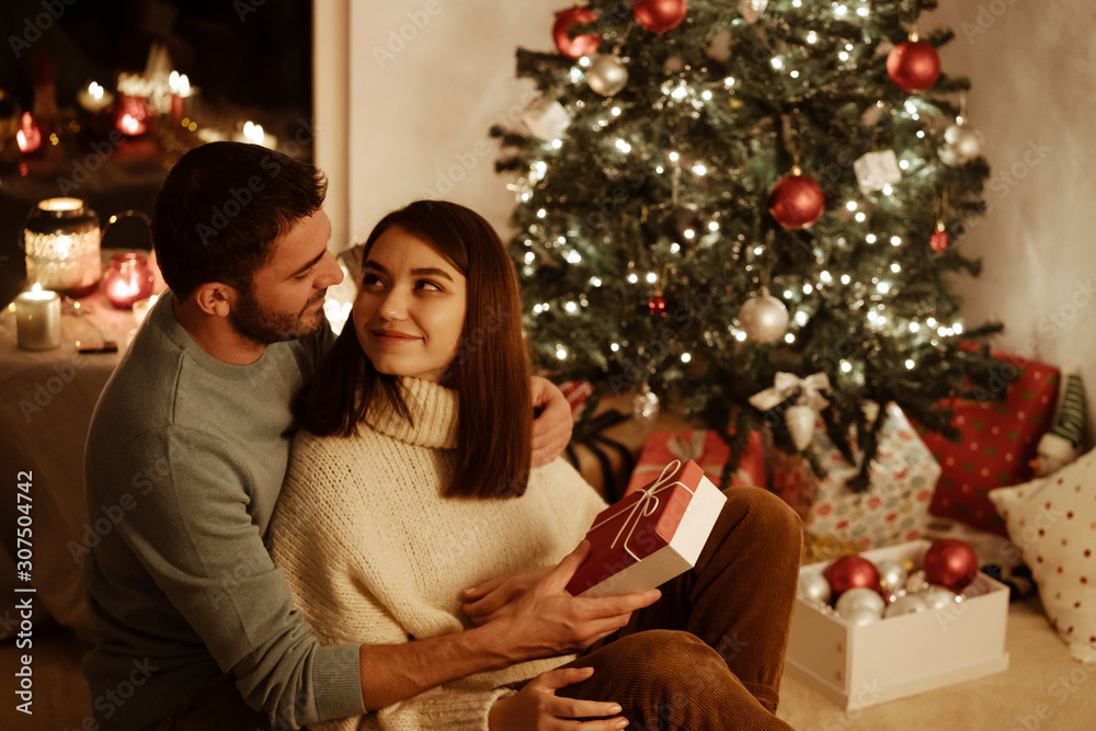 happy couple sitting near xmas decoration on background; man giving gift box to woman and they look at each other