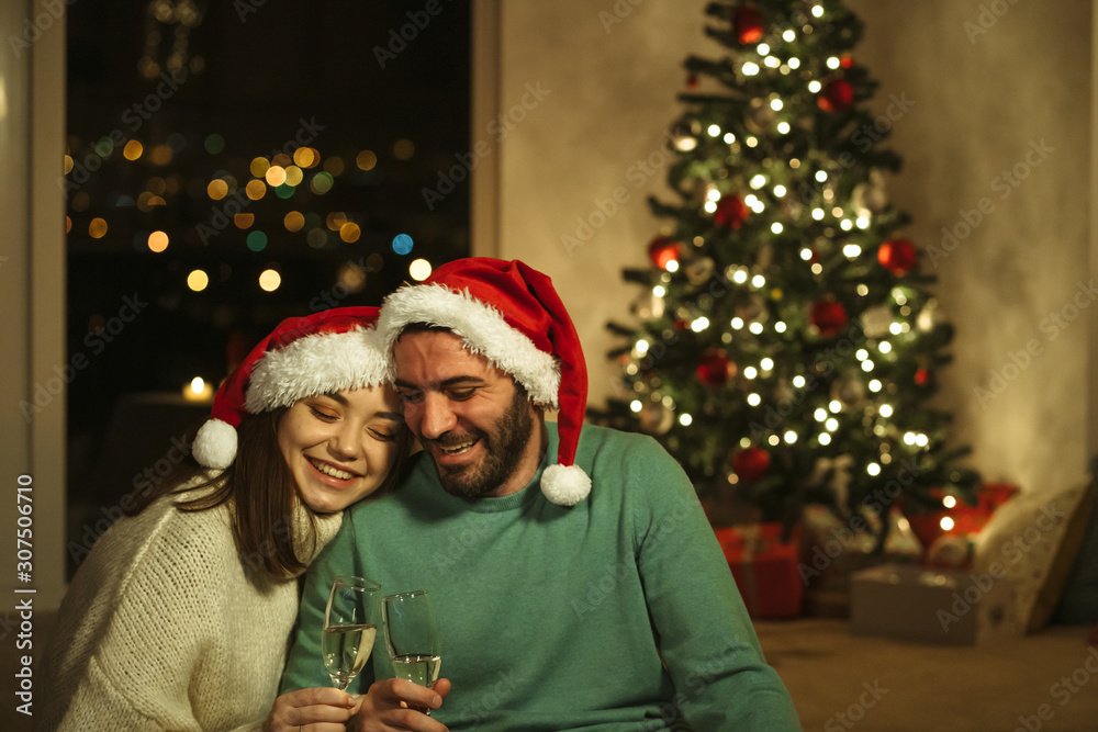 happy couple drinking champagne with santa hat and x-mas tree on background; christmas concept happy couple drinking champagne with santa hat and x-mas tree on background; christmas concept
