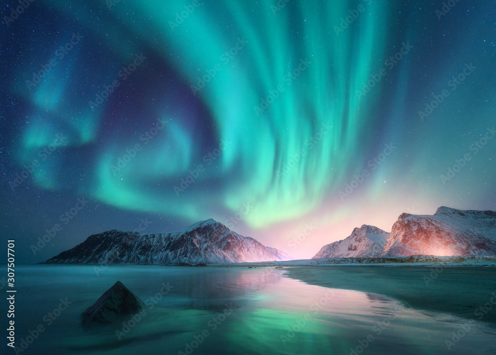 Aurora borealis over the sea and snowy mountains. Northern lights in Lofoten islands, Norway. Sky with polar lights and stars. Winter landscape with aurora, reflection, sandy beach at starry night