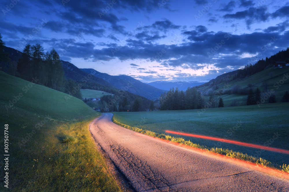Blurred car headlights on rural road at night in summer. Landscape with asphalt road, light trails, mountains, green grass, trees, and blue sky with clouds at dusk. Roadway through the meadows