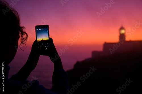 Portugal, Algarve, Over shoulder view of person taking smart phone photos of Cape Saint Vincent lighthouse at dawn