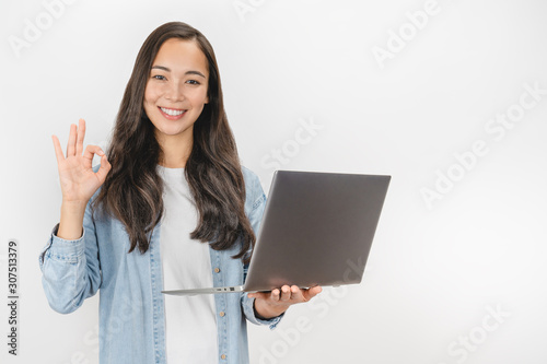 Portrait of young asian girl holding laptop computer and showing ok gesture isolated over white background