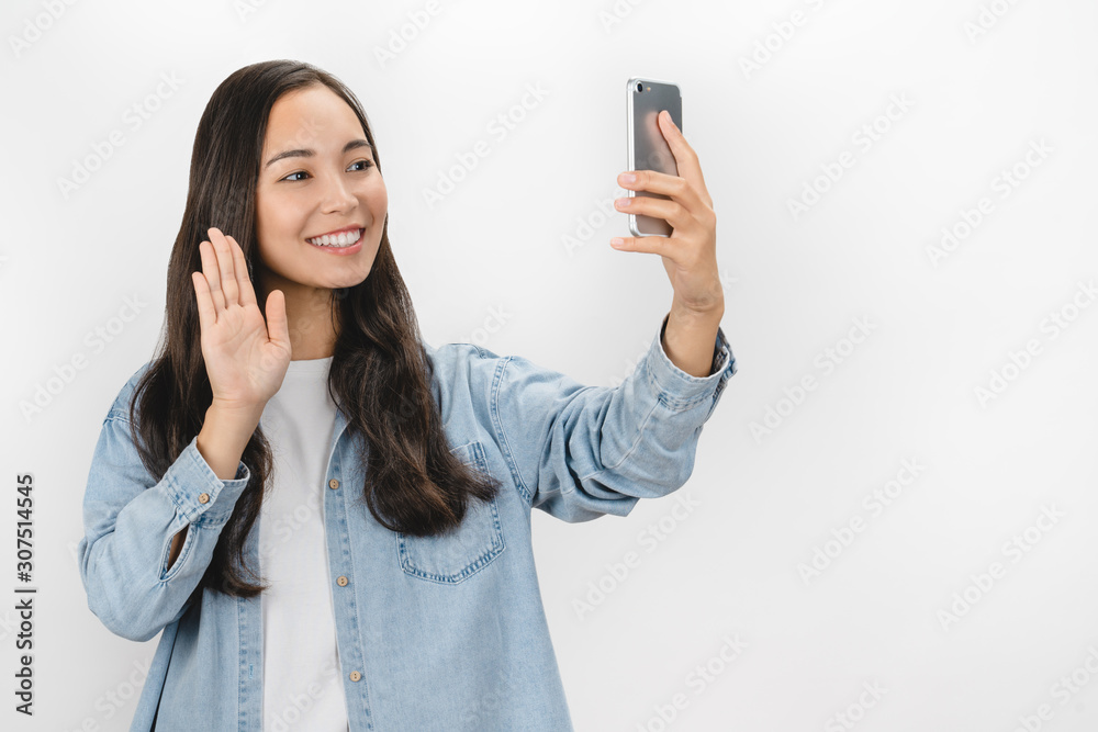 Portrait of adorable brunette asian woman using cellphone showing Hi gesture while making video call over white background Portrait of adorable brunette asian woman using cellphone showing Hi gesture while making video call over white background