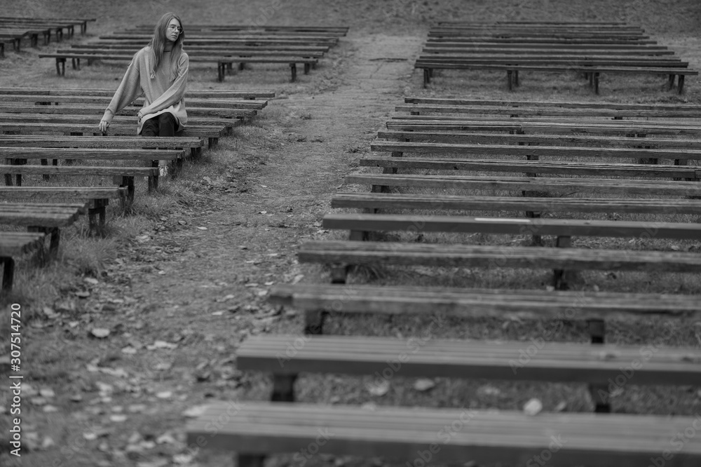 Obraz premium Sad depressed girl sitting alone on bench in park with multiple desolate benches around her. Loneliness and despair concept.