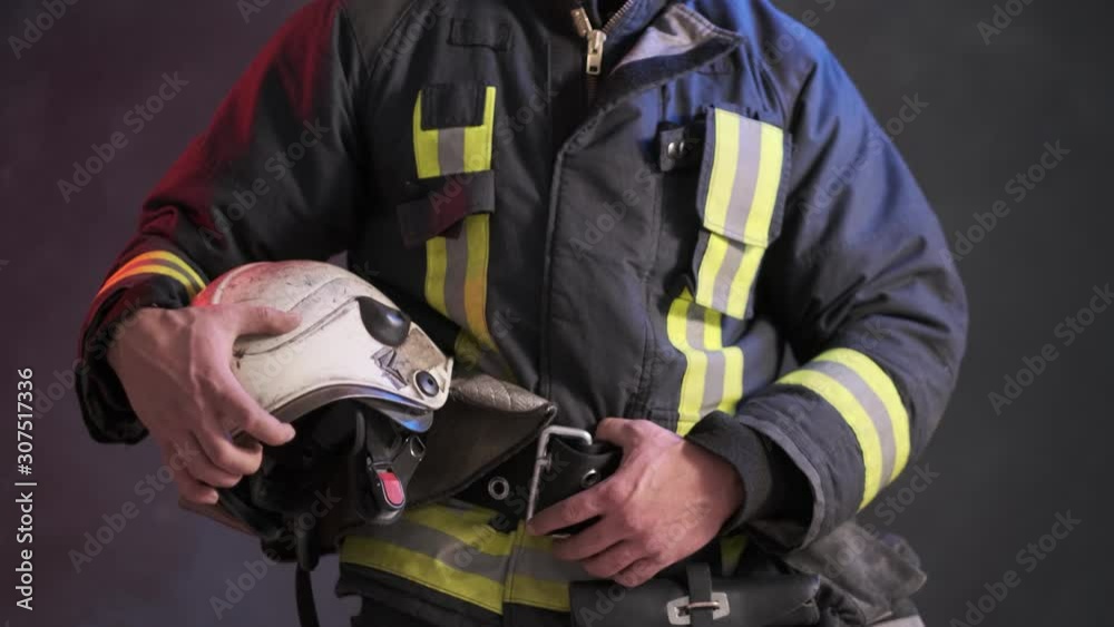 Close-up view of a brave firefighter in special uniform holds protective helmet standing against a gray wall Close-up view of a brave firefighter in special uniform holds protective helmet standing against a gray wall
