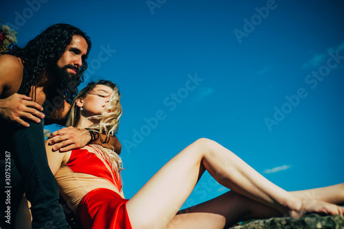 Couple in love. Strong handsome bearded man embracing his young sexy woman in elegant red dress at clear summer blue sky background. Couple enjoying their strong relations and love.