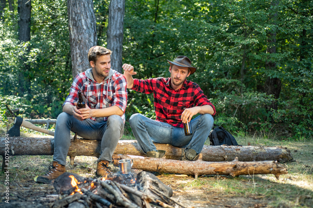 Happy young people camping in woods. Summer lifestyle. Young guy having a picnic. Enjoying camping holiday in countryside. Two Happy people sitting around campfire with beer. Happy young people camping in woods. Summer lifestyle. Young guy having a picnic. Enjoying camping holiday in countryside. Two Happy people sitting around campfire with beer.
