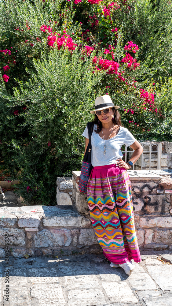 Asian woman in white t-shirt  in Athens in white hat and sarong leaning on stone wall in front of church