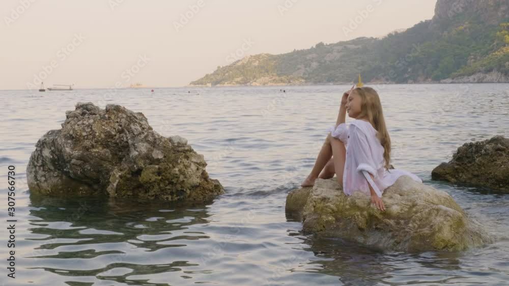 Relaxed girl in white tunic enjoying sea landscape on stone in water. Happy young girl sitting on rock in sea water on sunny beach at summer holiday Relaxed girl in white tunic enjoying sea landscape on stone in water. Happy young girl sitting on rock in sea water on sunny beach at summer holiday