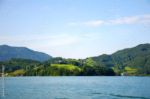Houses on a hill by the lake. Mountains with forest in the background. Beautiful clear sky, summer day. © Мария Аввакумова