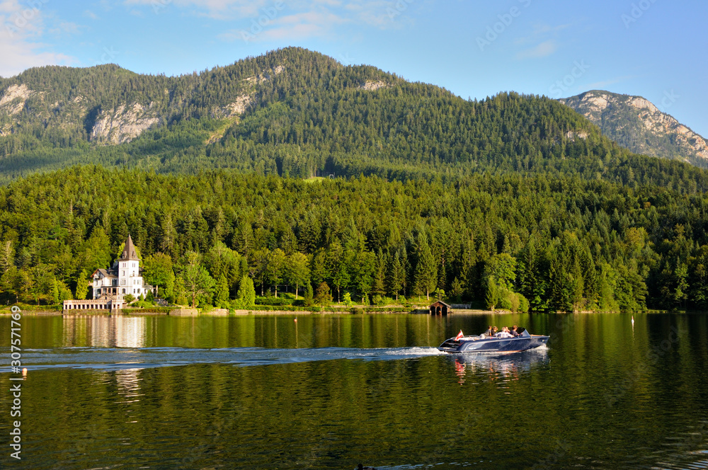 A large white house in the form of a castle on the shore of the lake. Green mountains in the background. Motor boat on the lake.