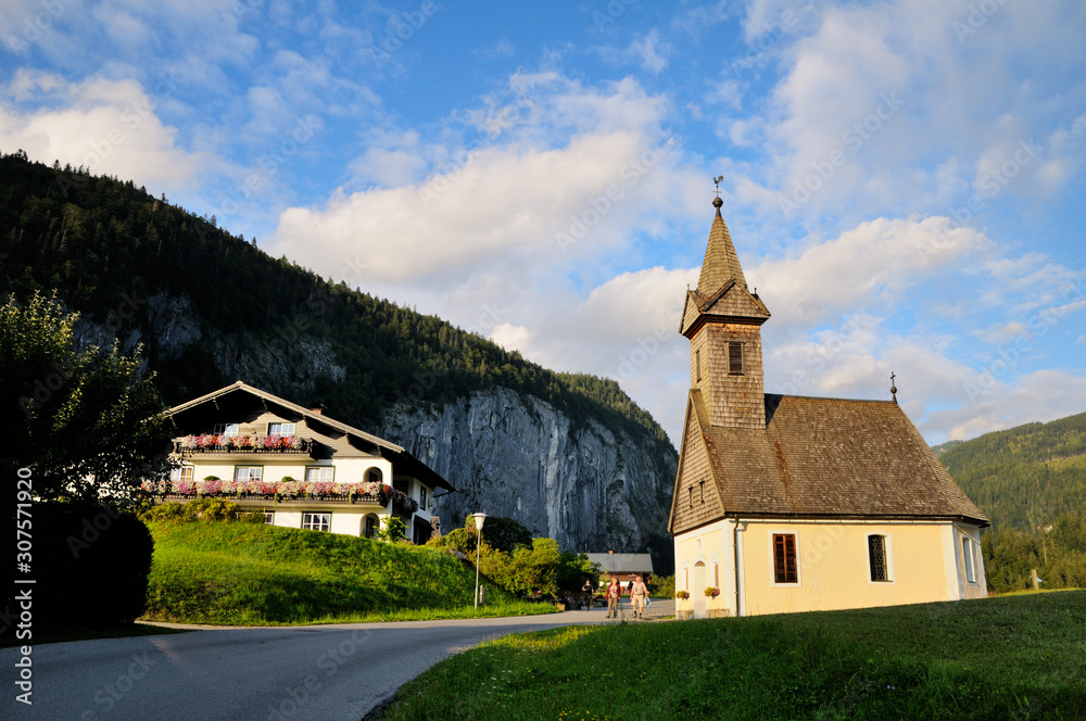 A small Catholic church with a wooden roof in a Bavarian village. Beautiful evening lighting. Blue sky and mountains in the background.