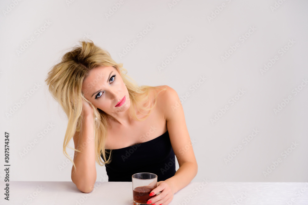 Portrait of a pretty blonde model woman with excellent make-up, beautiful hair and clean skin on a white background sitting at a table with a glass of whiskey in her hands. Portrait of a pretty blonde model woman with excellent make-up, beautiful hair and clean skin on a white background sitting at a table with a glass of whiskey in her hands.
