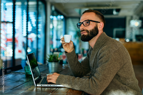 Young bearded man with glasses in a cafe working on laptop and having espresso coffee	