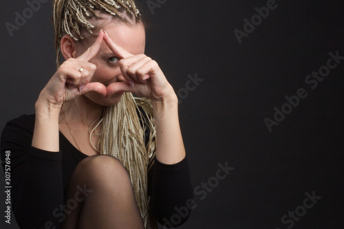 Obraz na płótnie Studio portrait of a girl with dreadlocks afro hairstyle Zizi on a gray background holds fingers in the form of a triangle and looks through them to the camera in dark clothes w salonie