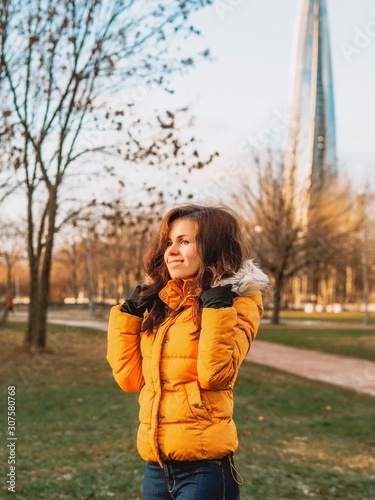 A young girl with long hair in a yellow winter jacket stands in the Park at the beginning of winter against the backdrop of a skyscraper