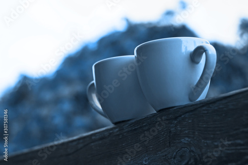 Two cups of coffee in classic blue color of the Year 2020 on wooden balcony on the background of sunset with bokeh light